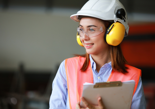 Técnica em segurança do trabalho com EPI, capacete, óculos e protetor auricular durante inspeção industrial.